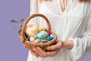 Young woman holding basket with makeup products, eucalyptus and Easter decor on lilac background - Powered by Adobe