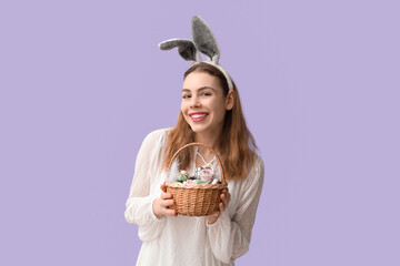 Happy smiling young woman in bunny ears headband holding basket with makeup products, flowers and Easter decor on lilac background - obrazy, fototapety, plakaty