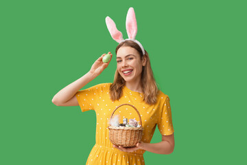 Happy smiling young woman in bunny ears headband holding basket with makeup products, flowers and Easter egg on green background - obrazy, fototapety, plakaty