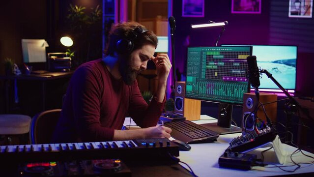 Musician artist taking notes composing new song in his home studio, writing down lyrics and musical notes before recording his melody. Songwriter producing music with daw software tools. Camera B.
