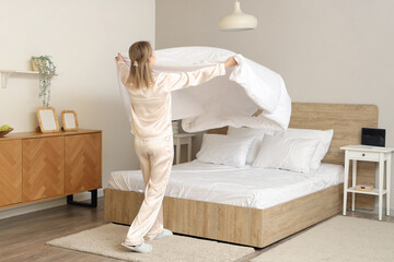 Pretty young woman in pajamas making bed in light bedroom