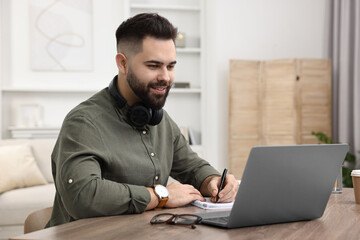 E-learning. Young man taking notes during online lesson at wooden table indoors