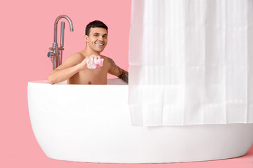 Young man in bathtub with loofah and curtain on pink background