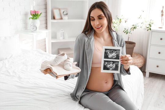 Young pregnant woman with baby clothes and sonogram image sitting in bedroom