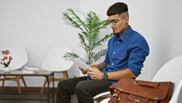 Young latin man with a tattoo, engrossed in business paperwork, anxiously checking the clock in a waiting room.