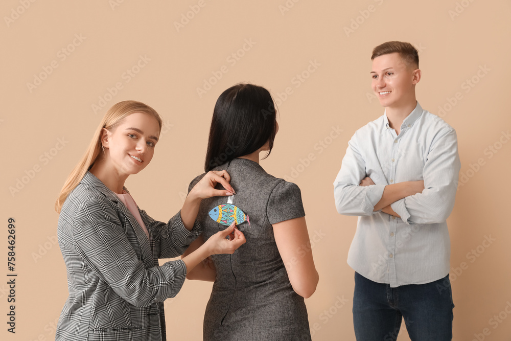 Sticker Young woman sticking paper fish on her colleague's back against beige background. April Fools' Day celebration