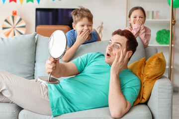 Little kids laughing at their shocked father with paintings on his face at home. April Fool's Day...