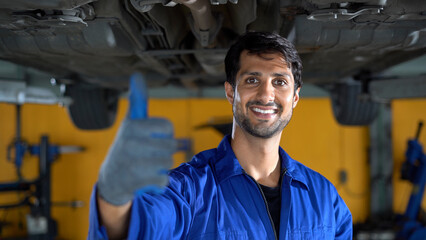 happy latin auto mechanic man checking tires , brakes under car in garage cars service . hispanic technician repairing vehicle show thumbs up and looking camera . lifted car checklist tyres and wheel - obrazy, fototapety, plakaty