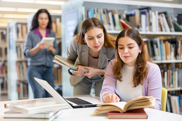 Two female students studying at the university are preparing for the exam in the library on a laptop looking for information ..in textbooks