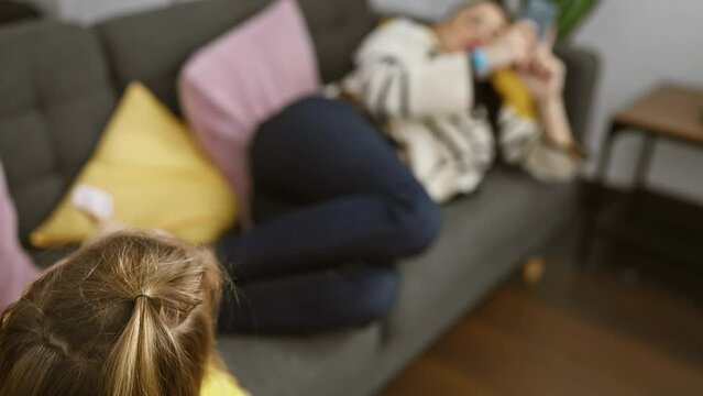 A mother relaxes on the couch using her smartphone while her young daughter plays beside her in a cozy living room