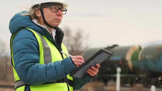 Attractive female inspector in white helmet and green vest, despite windy weather, makes report on the shipment of wagons using a digital tablet and stylus, against background of train with petroleum 