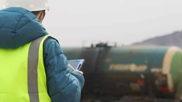 Female inspector in hardhat and vest, makes report on shipment of wagons using tablet and stylus, against background of train with oil, view from back, mid shot.