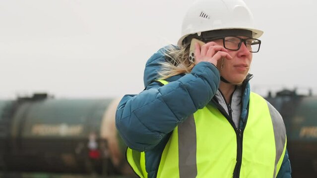 Portrait of woman constructor wearing helmet and safety vest, monitoring the departure of a freight train giving instructions by phone against the backdrop of a standing carriage.