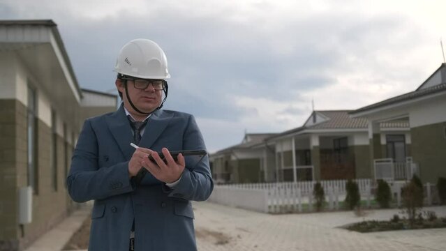 Portrait of male engineer technologist in white helmet with clipboard, standing among new buildings. Man realtor in protective hard hat and suit.
