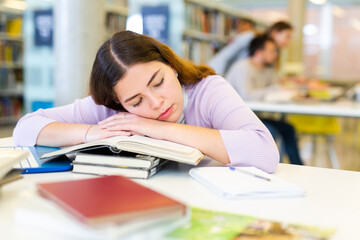 Tired young woman lying on stack of books in public library