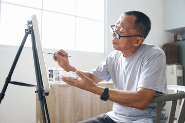 Elderly Asian man wear glasses painting at canvas at home