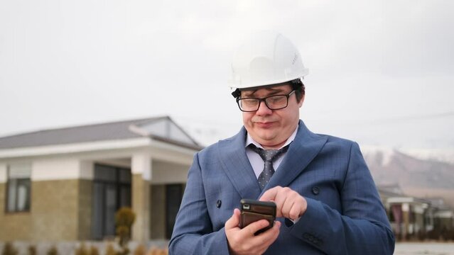 Boss in suit and ties with protective white helmet on head, dissatisfied with result of construction work, typing message on phone using touchscreen while standing against backdrop of an suburban area