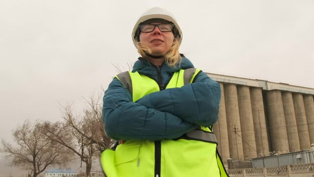 Young woman contractor in protective hard hat with vest stands with crossed arms on street next to factory accept work.
