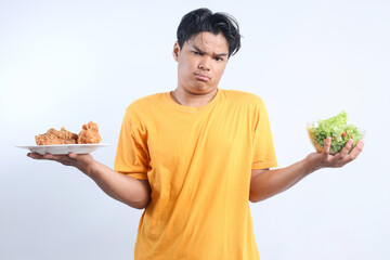 Young Asian man holding a plate of fried chicken and a bowl of salad, confuse to deciding which one to eat. Healthy food and unhealthy food concept