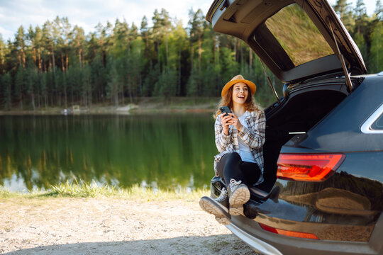 Happy woman  with a phone in her hand sits in the trunk of a car. Concept of technology, adventure, travel by car.