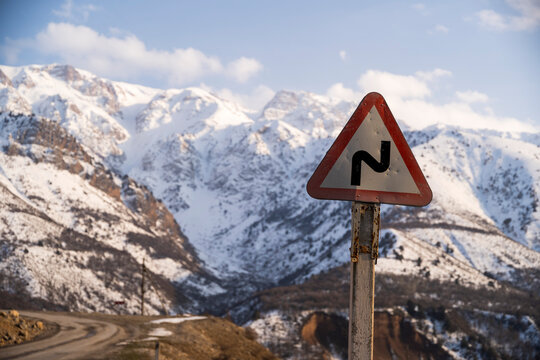 A winding mountain road disappears into the distance, framed by a cautionary road sign and the backdrop of snow-clad mountains. This image speaks to the careful attention needed when navigating such t