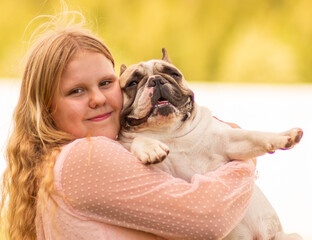 On a hot summer day outside, a beautiful, elegant girl is holding a French bulldog in her arms. A...