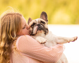 On a hot summer day outside, a beautiful, elegant girl is holding a French bulldog in her arms. A...