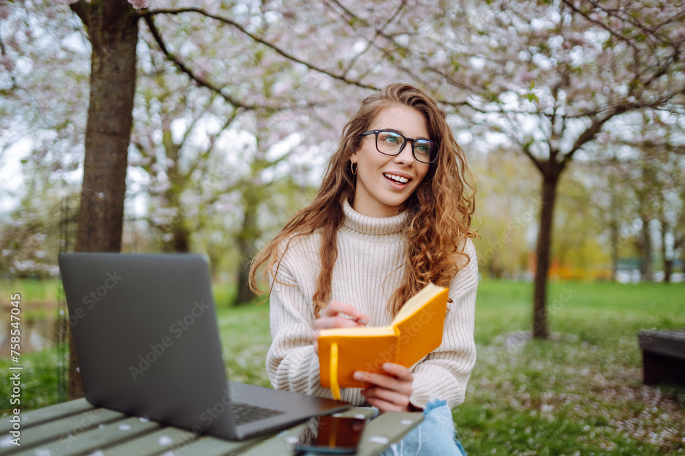 Wall mural positive student with laptop reading book while doing homework assignment on green meadow in campus 