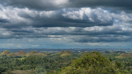 Many rounded orange-brown hills stretch to the horizon. Lush tropical green vegetation in the valley. Clouds in the blue sky. Chocolate Hills Natural Monument. Philippines. Bohol Island