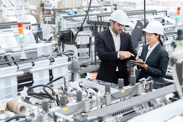 An American male engineer and an Asian female company owner in suits and hard hats use laptops to...