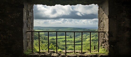 A stunning view of a lush green valley framed by a stone window with a metal railing, overlooking a grassy landscape under a clear sky with fluffy cumulus clouds