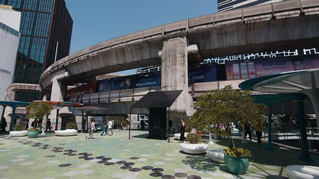 bustling public space in Bangkok, featuring pedestrians, an elevated skytrain, contemporary architecture, and decorative planters under a clear blue sky