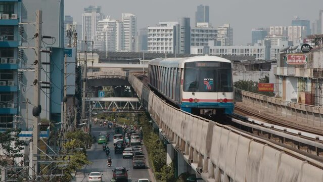 A sky train travels along its elevated track above a busy road with motorbikes and cars in Bangkok, with a backdrop of urban buildings and slightly hazy sky.