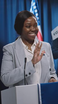 Medium vertical shot of young African American female Democratic nominee running for president or Senate walking on tribune, smiling, waving, greeting public, before making policy statement
