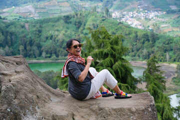 Woman enjoying windy atmosphere, sitting on a rock over the hill facing calm lake in the distance