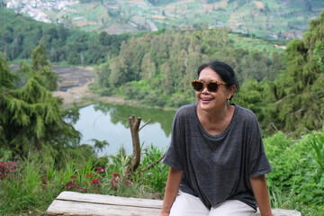 Asian female tourist sitting on a bench at a breezy hilltop with beautiful lake in the background