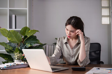 Unhappy Asian businesswoman shows stress while working in a home office decorated with soothing green plants.