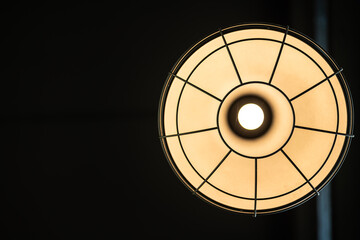 A classic style round ceiling lamp with glowing lightbulb in warm light shade, with dark area as background. Interior decoration and object photo, close-up.