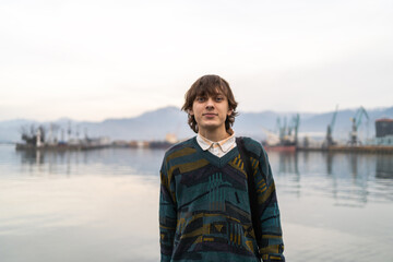 Young man in vintage inspired attire stands before tranquil harbor backdrop, fusing classic style with maritime setting.