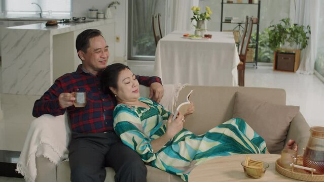 Wide shot of aged Asian spouses talking while resting on couch on weekend at home