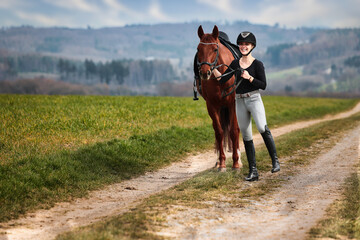 Rider and horse stand side by side on a path in a wide spring landscape.