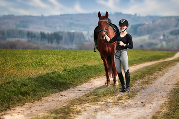 Rider and horse stand side by side on a path in a wide spring landscape.