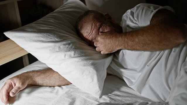 Handsome middle-aged caucasian man with a beard, tired and stressed, lying on the bed in a dimly lit bedroom, revealing a portrait of life&rsquo;s worries