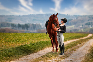 Rider and horse stand side by side on a path in a wide spring landscape.