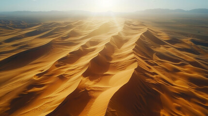 Aerial view of vast desert dunes under a bright sunrise, showcasing the textured patterns of sand and the play of light and shadows.