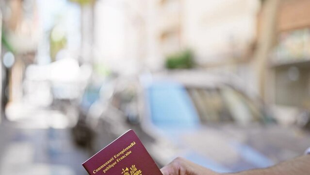 Hand holding a french passport outdoors in a city environment, with blurred urban street scenery in the background.
