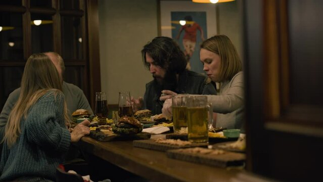 Group of friends talk, eat burgers, snacks and drink beer sitting in stylish bar or pub together in the evening. People having dinner, resting in restaurant on weekend. Lifestyle and leisure concept.