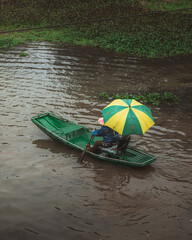 A lady rows a boat in a river covered by a green and yellow umbrella
