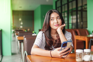 Thoughtful young Asian woman wearing dress sitting at a restaurant for a breakfast, holding a smartphone while propping her chin - obrazy, fototapety, plakaty