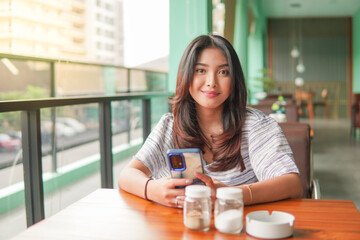 Smiling young Asian woman wearing dress and feeling happy, sitting at a restaurant for a breakfast while holding a smartphone